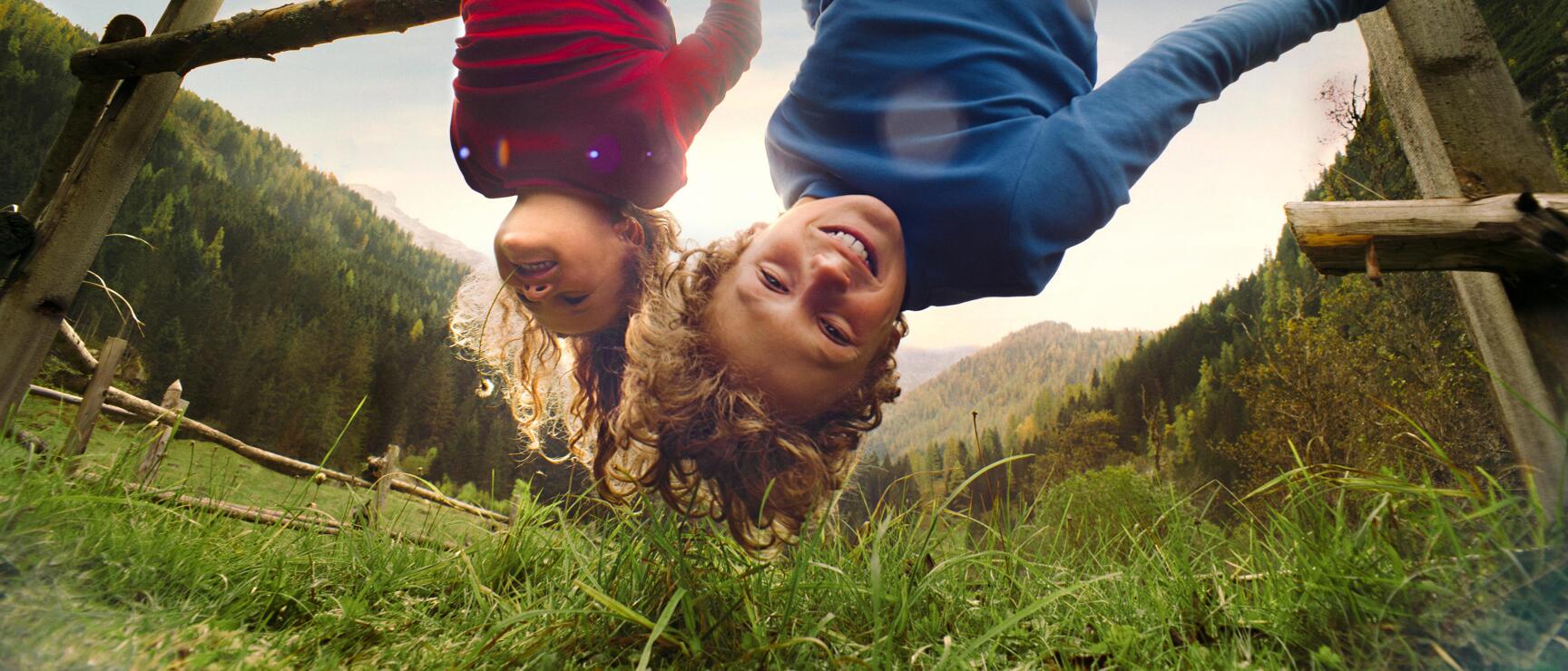 Zwei Kinder hängen kopfüber lachend an einem Holzzaun, grüne Almwiese und Bergwald im Hintergrund.