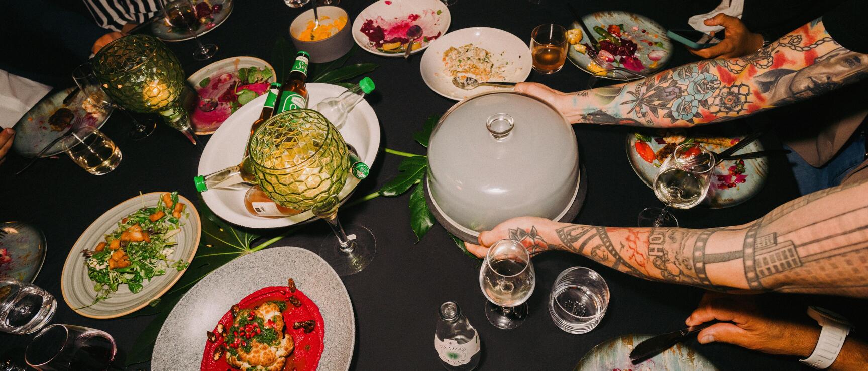 Gedeckter Tisch mit bunten Speisen, Weingläsern und tätowierten Armen beim Servieren.