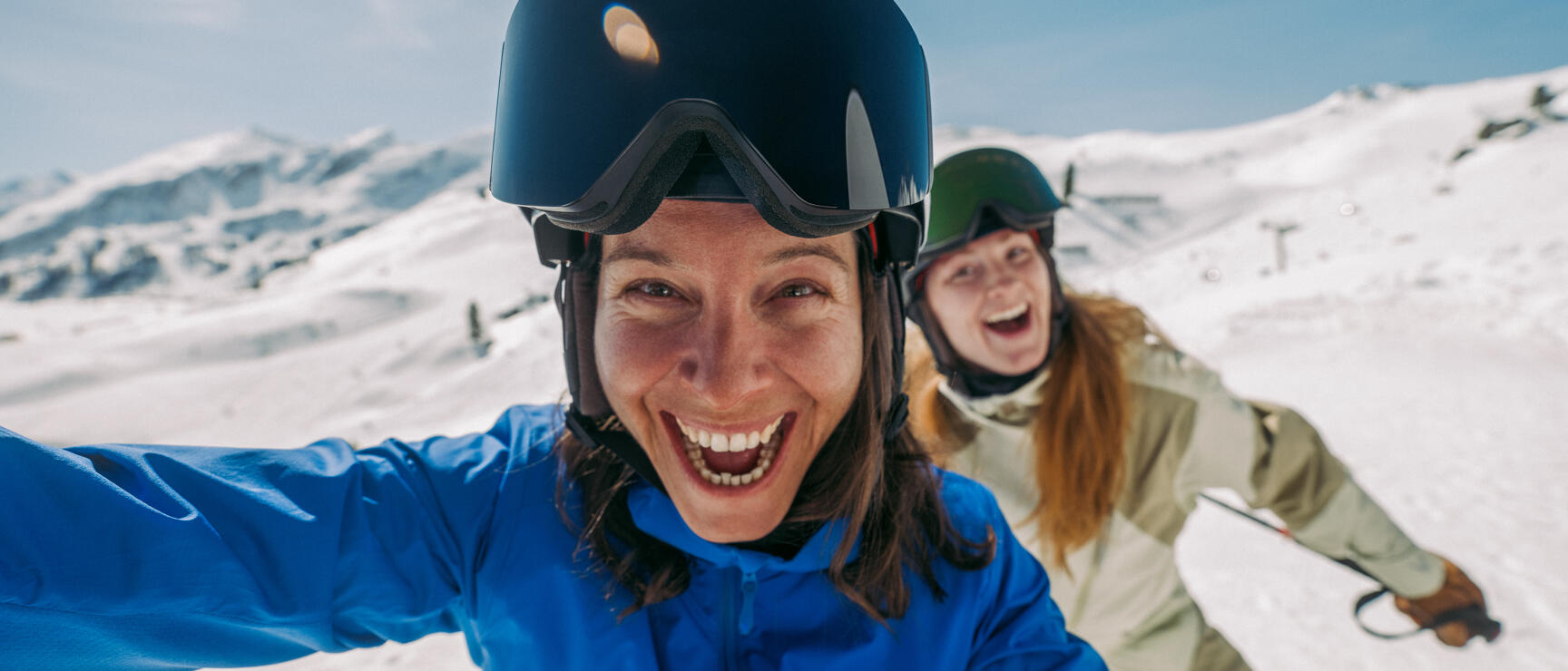 Zwei lachende Skifahrerinnen mit Helm und Skibrille machen ein Selfie auf einer verschneiten Piste.