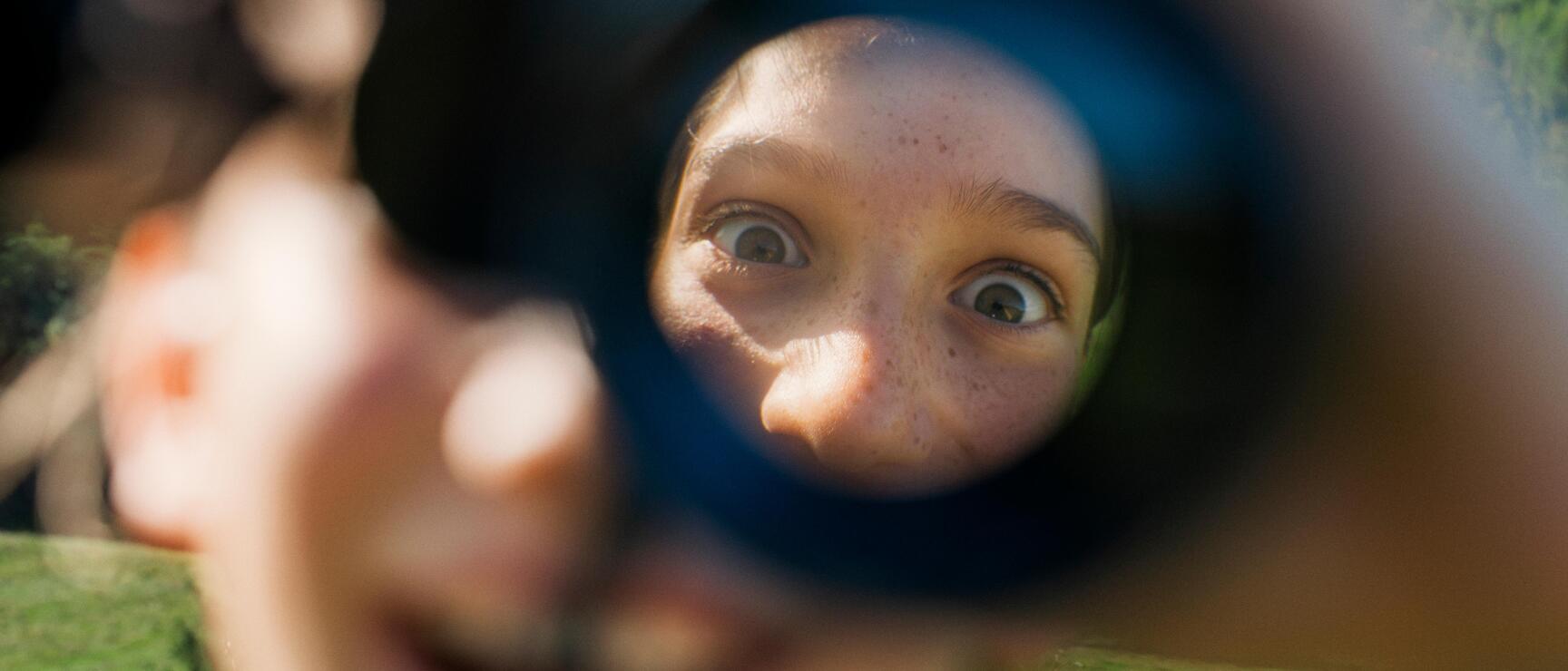 Ein Kind blickt neugierig durch ein Fernglas, Nahaufnahme mit unscharfem Vordergrund, grüne Almwiese und Wald im Hintergrund