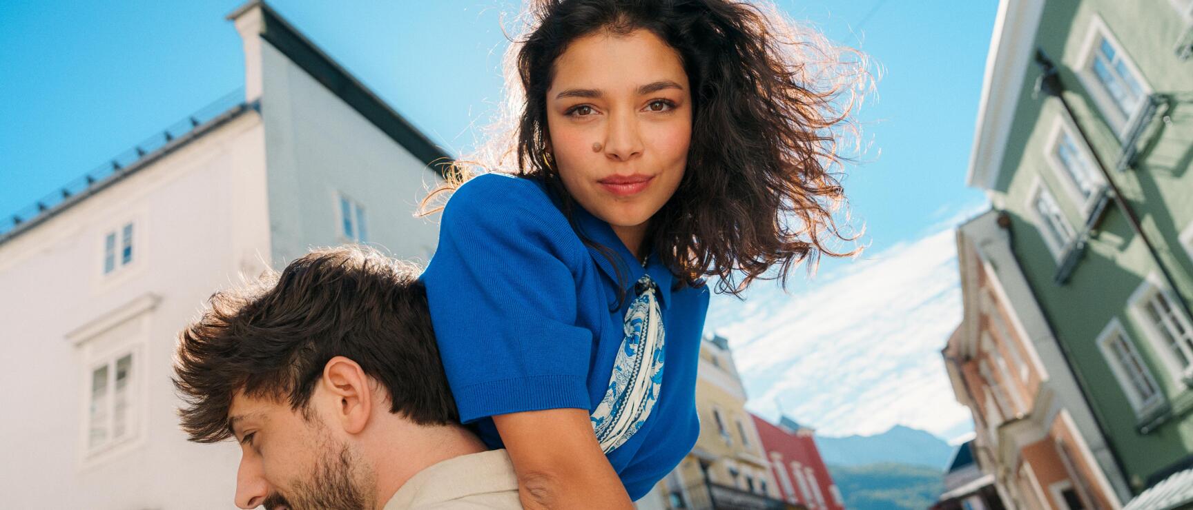 Woman in blue shirt on man's back with colourful row of houses and mountains in background.