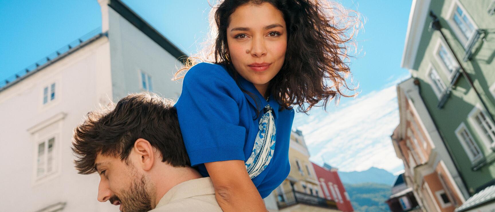 Frau im blauen Shirt auf dem Rücken eines Mannes, bunte Häuserzeile und Berge im Hintergrund.