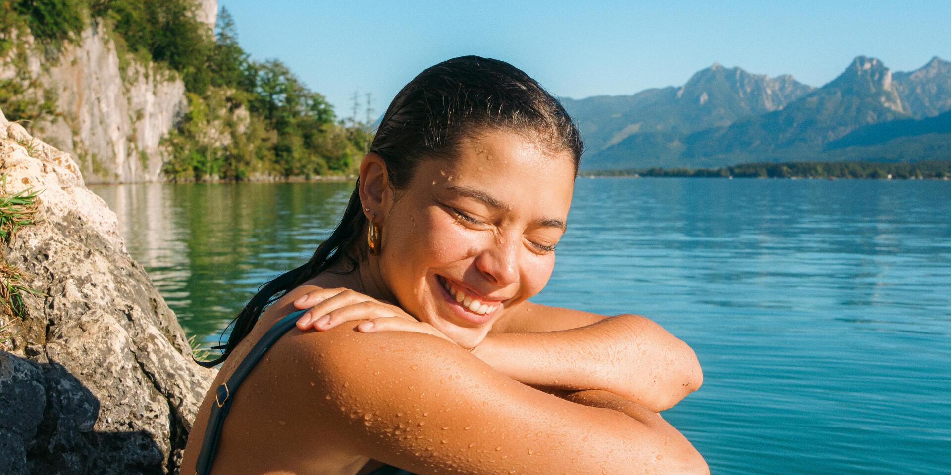 Lebensgefühl Österreich - Kampagnenbild Eine Frau sitzt nach dem Schwimmen auf einem Felsen am See © Österreich Werbung / Marko Mestrovic Lachende Frau im nassen Badeanzug sitzt auf Felsen an einem Bergsee, Felswand und Berge im Hintergrund.