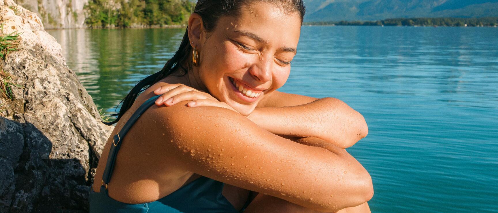 Eine Frau im Badeanzug sitzt lachend nach dem Schwimmen auf einem Felsen am Ufer eines Sees, mit Wassertropfen auf der Haut mit bewaldeter Felswand und Alpenpanorama im Hintergrund