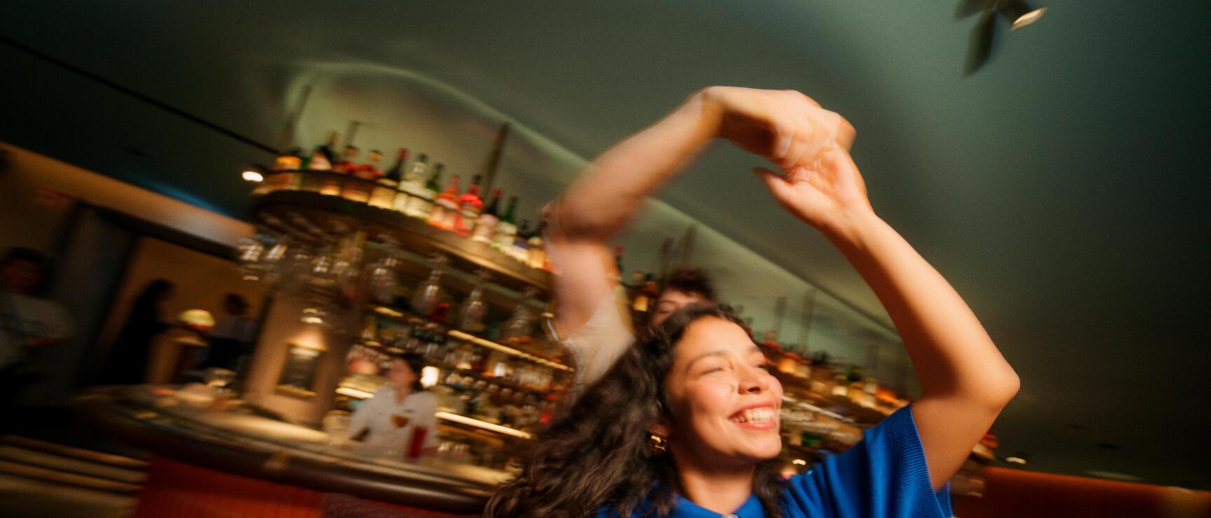 Eine Frau tanzt lachend mit erhobenen Armen in einer Bar, im Hintergrund eine beleuchtete Theke mit Flaschen, die Szene wirkt durch Bewegungsunschärfe lebendig