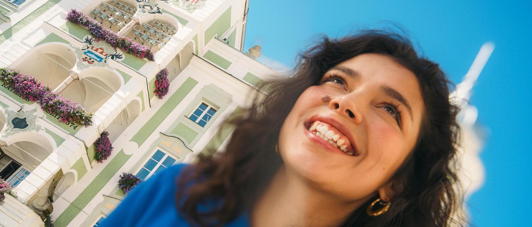 Lächelnde Frau vor dem grün-weißen Rathaus mit Blumenschmuck und Uhr in Gmunden, blauer Himmel.