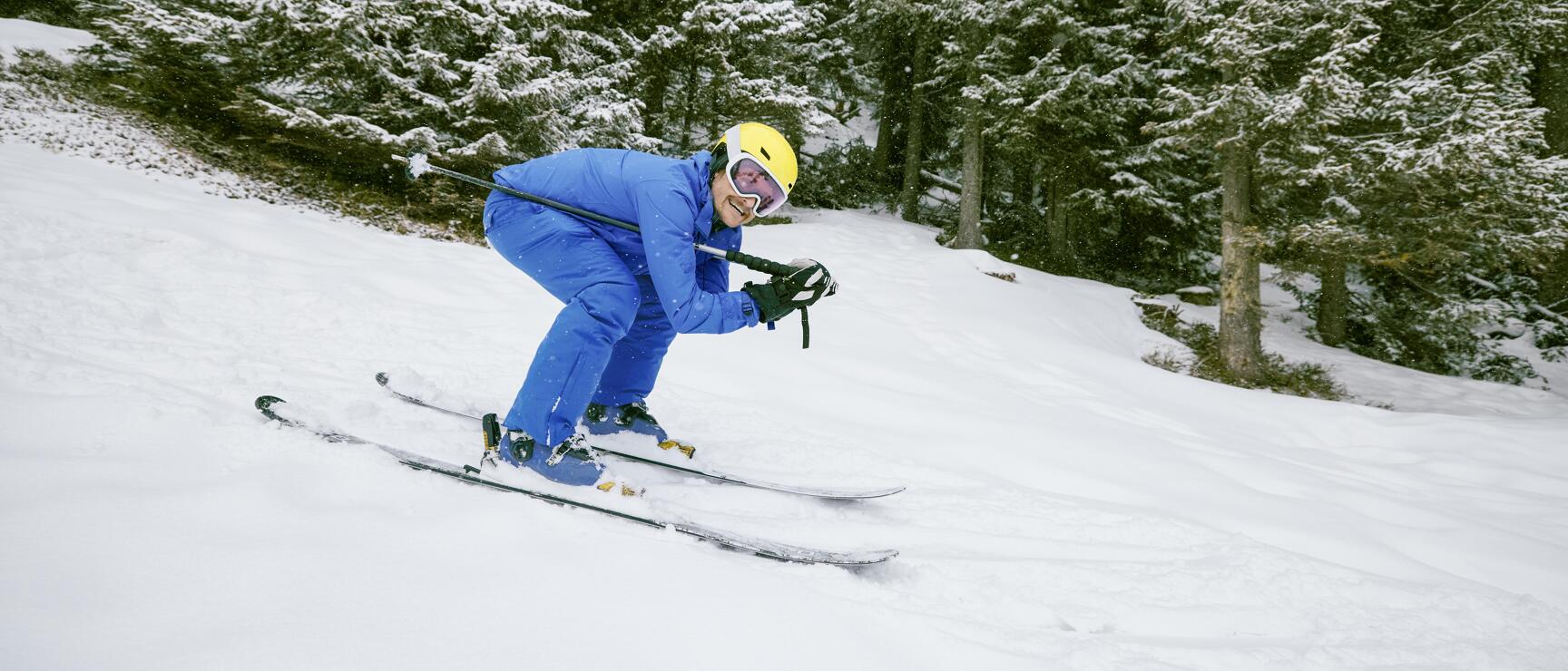 Skifahrer in blauem Anzug und gelbem Helm fährt Steilhang hinunter, verschneite Tannen im Hintergrund.