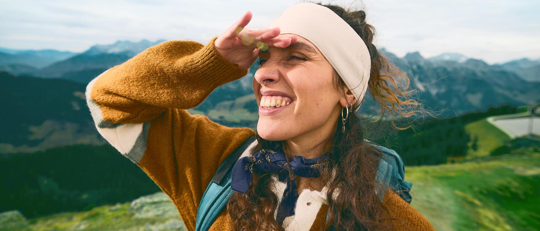 Lachende Frau mit Stirnband und Rucksack schirmt Augen ab, Bergpanorama im Hintergrund.