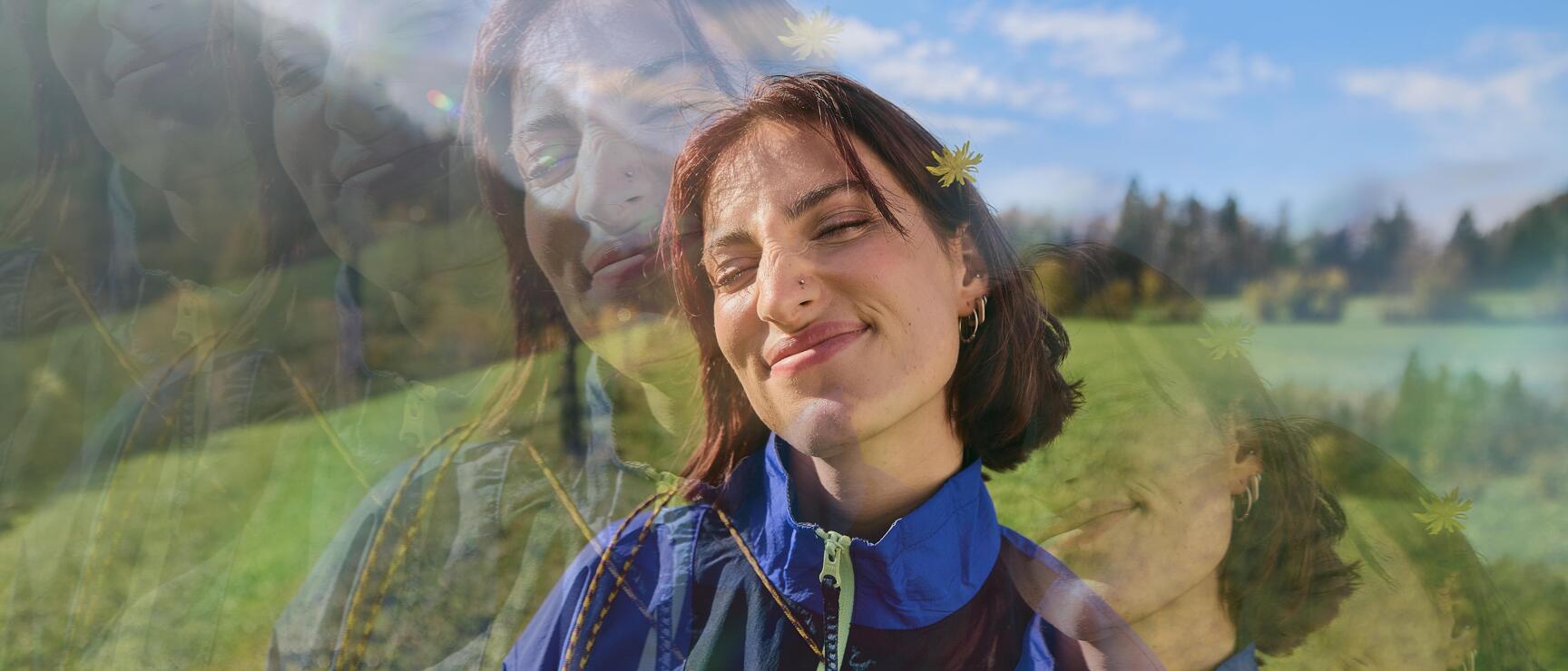 Junge Frau mit Blume im Haar und blauer Jacke auf grüner Wiese, blauer Himmel, Doppelbelichtung.