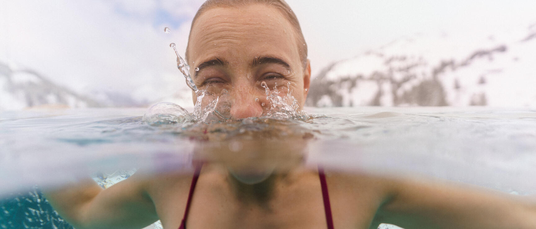 Woman in red bikini emerging from icy mountain lake with snow-covered peaks in background.