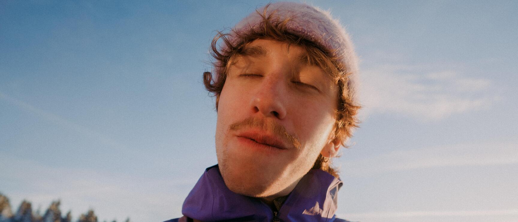 Mann mit rosa Mütze und lila Jacke isst im Freien, verschneite Bäume und blauer Himmel im Hintergrund.