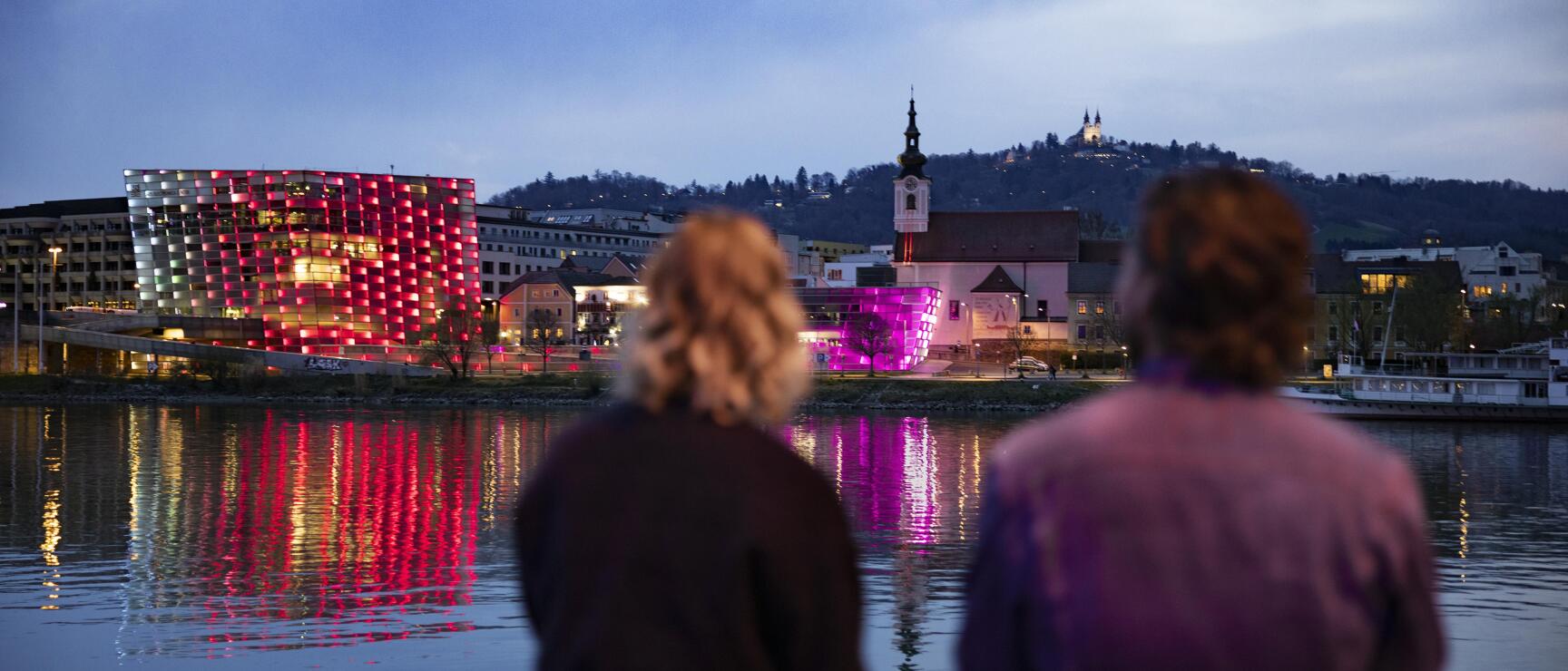 Two people looking from the Danube bank at the illuminated Ars Electronica Center in Linz at dusk.