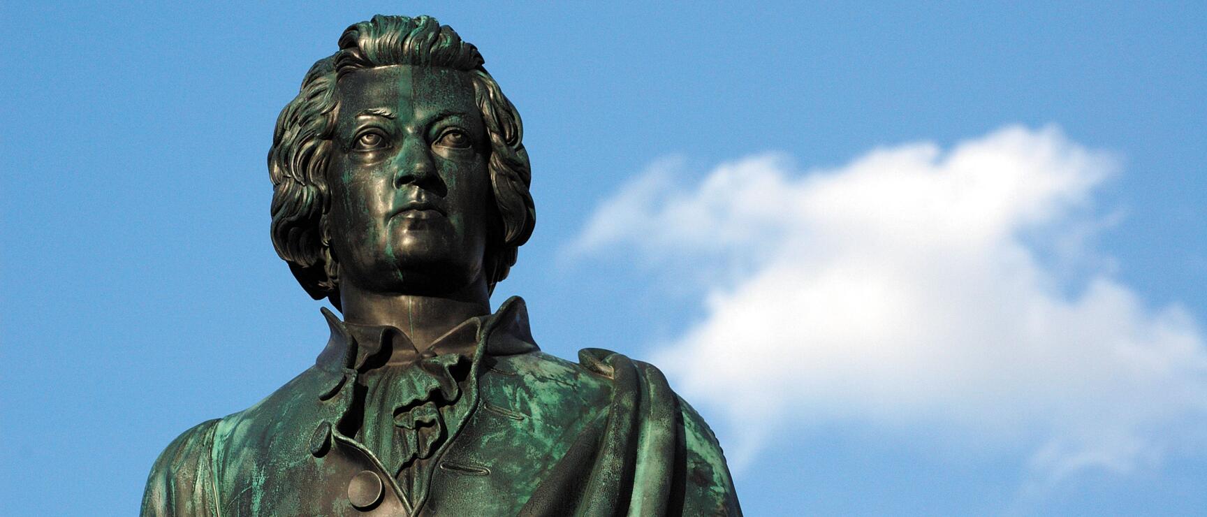 Bronze statue of a male figure with curly hair against blue sky with white cloud.