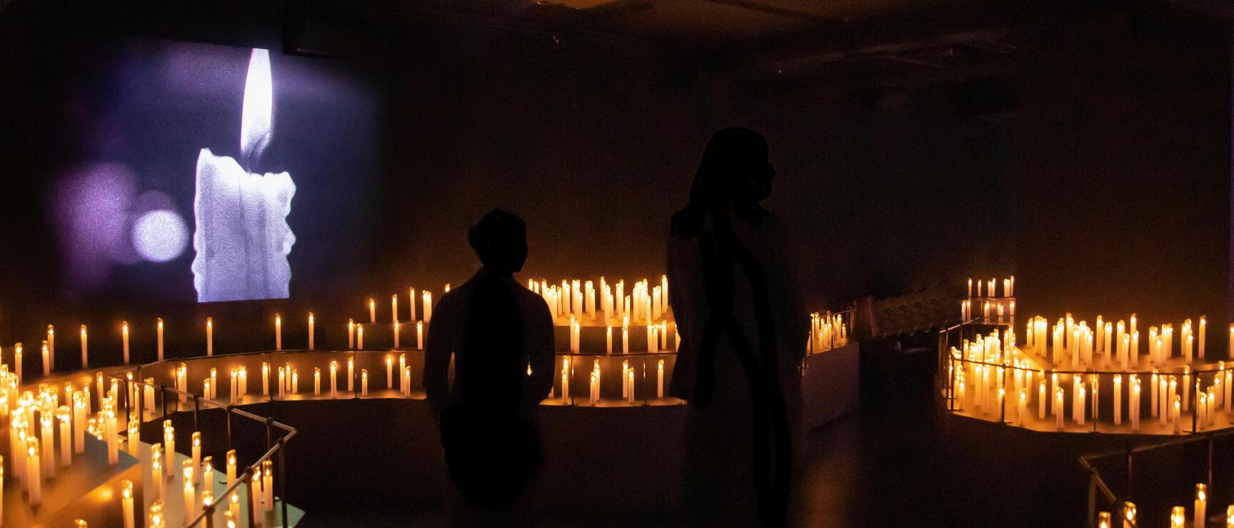 Two silhouettes in dark exhibition room surrounded by hundreds of candles, video projection of candle on wall