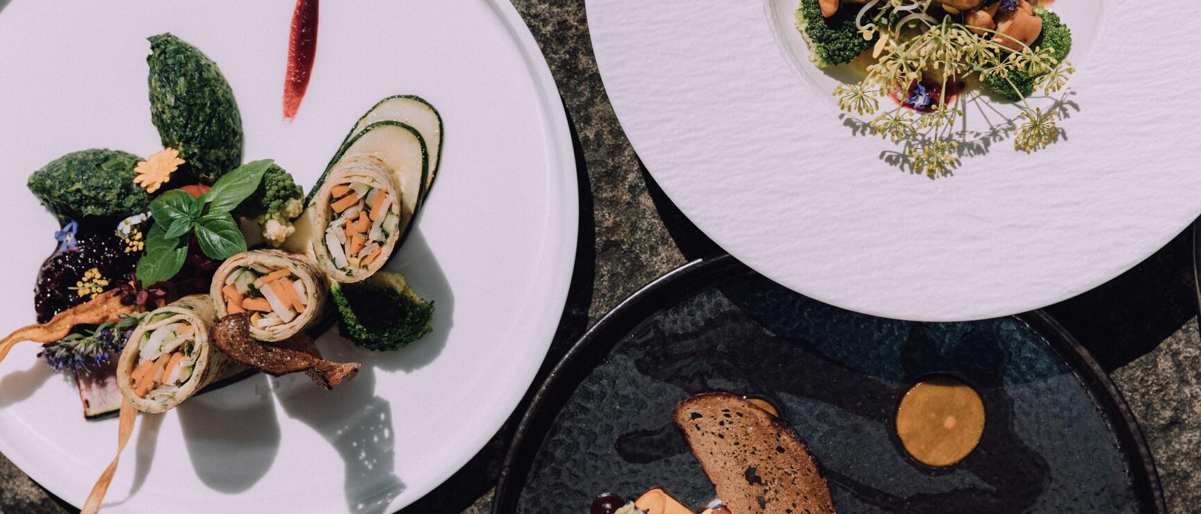 Three gourmet plates with artfully arranged vegetable dishes, flowers and herbs, overhead view.
