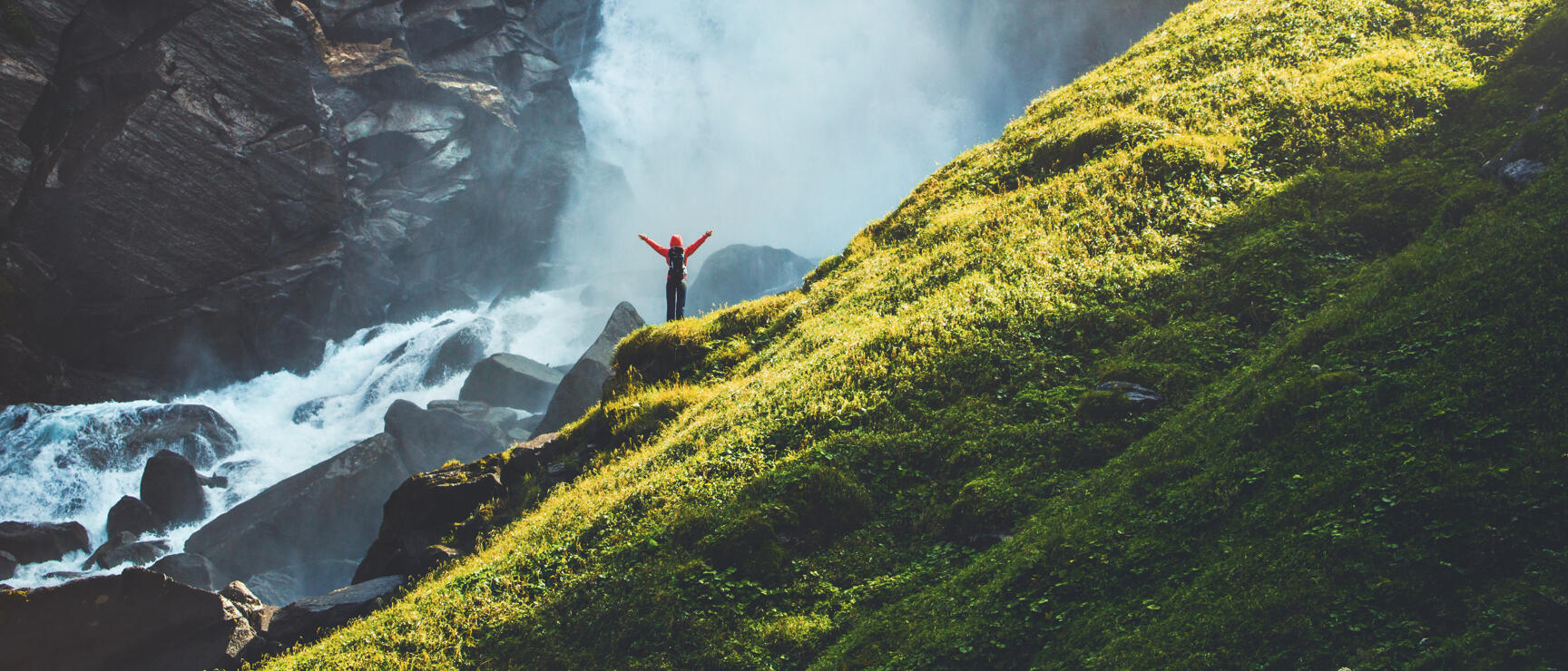 Person in red jacket with arms outstretched on rocks in front of large waterfall, green alpine meadow on right.