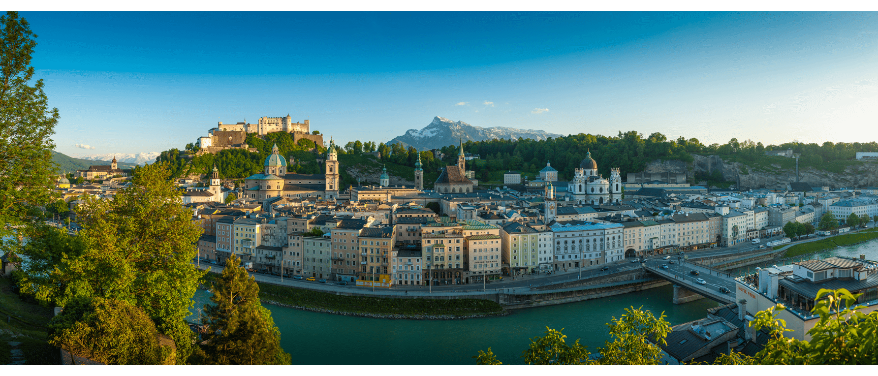 Panoramic view of Salzburg's old town with Hohensalzburg Fortress, cathedral, Salzach River and snow-capped peak in the background.