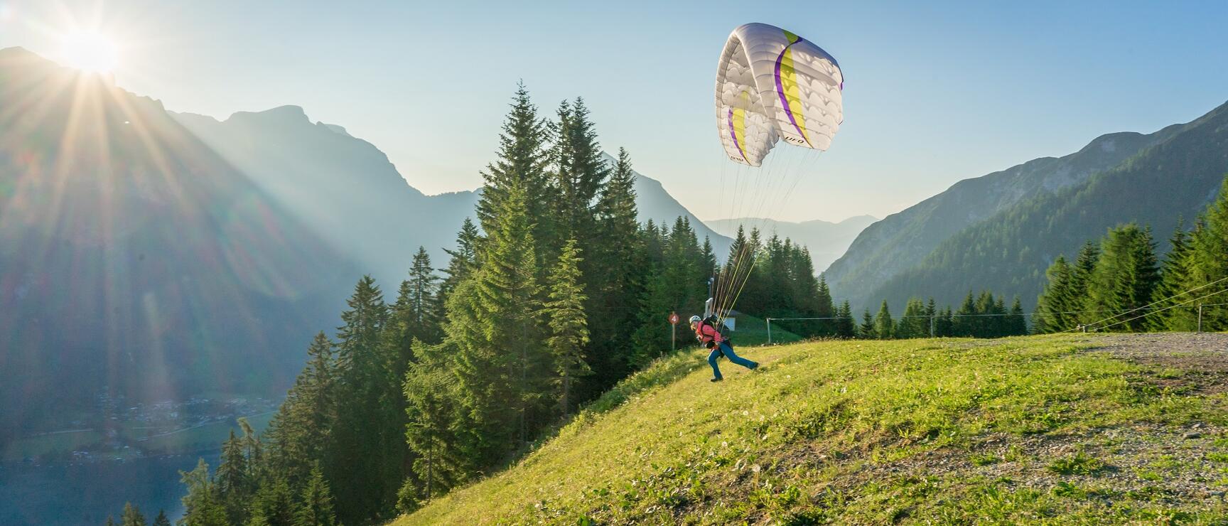 Person starting paraglide on green mountain meadow, coniferous forests and alpine panorama backlit.