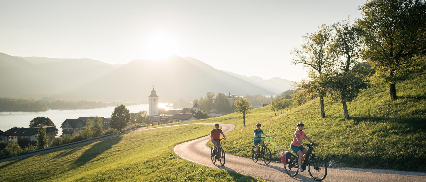Three cyclists on a winding path with church, lake and mountains backlit in the background.