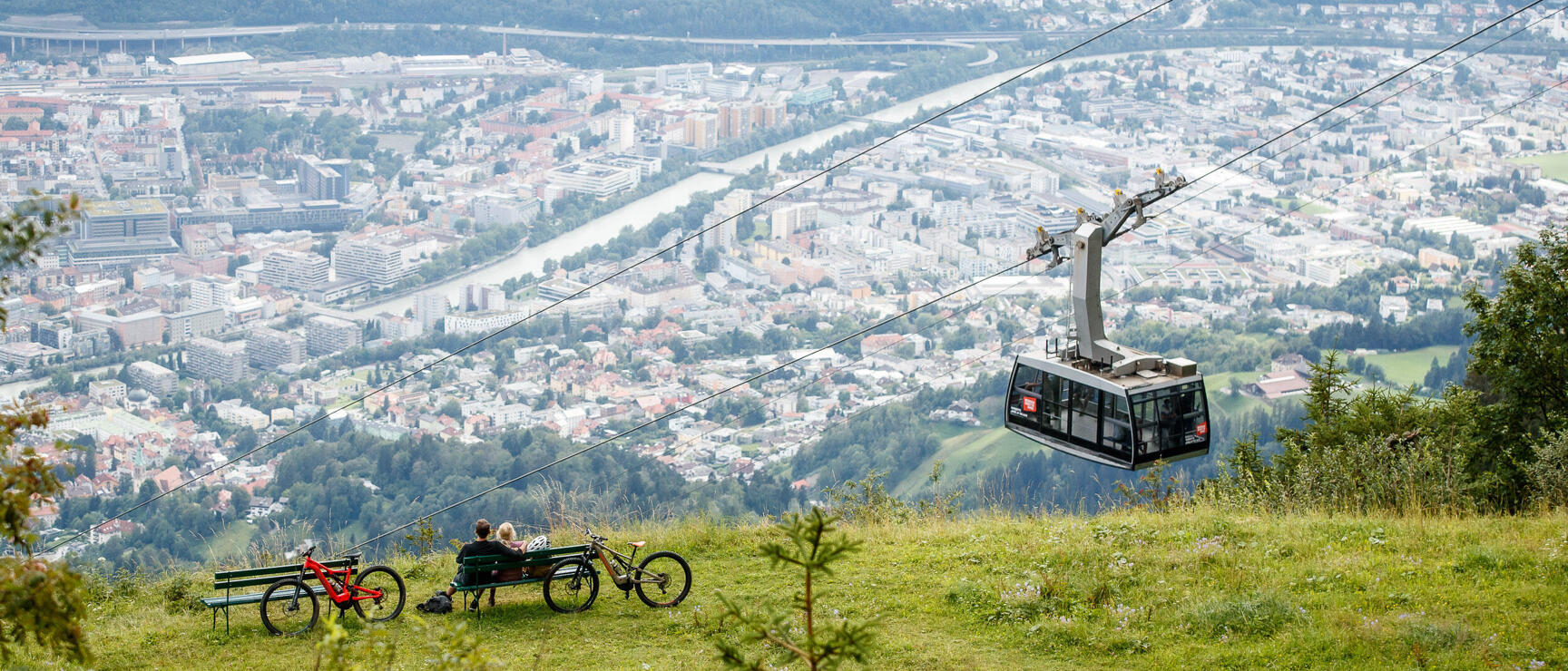 Two people with bicycles on a bench above Innsbruck, cable car cabin in the foreground.