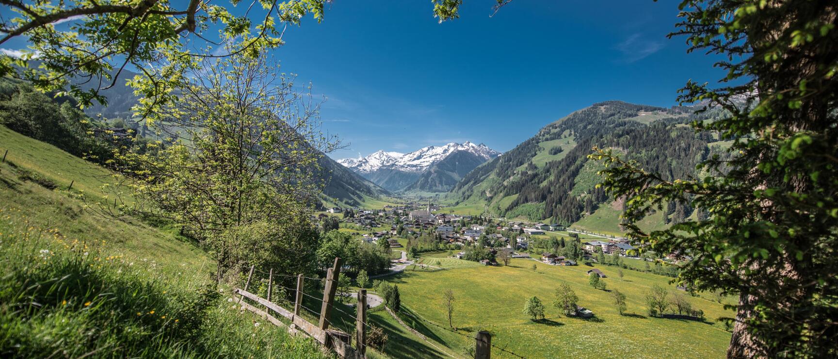 View of an alpine village in the valley with green meadows, wooden fence in foreground, snow-capped peaks in background.