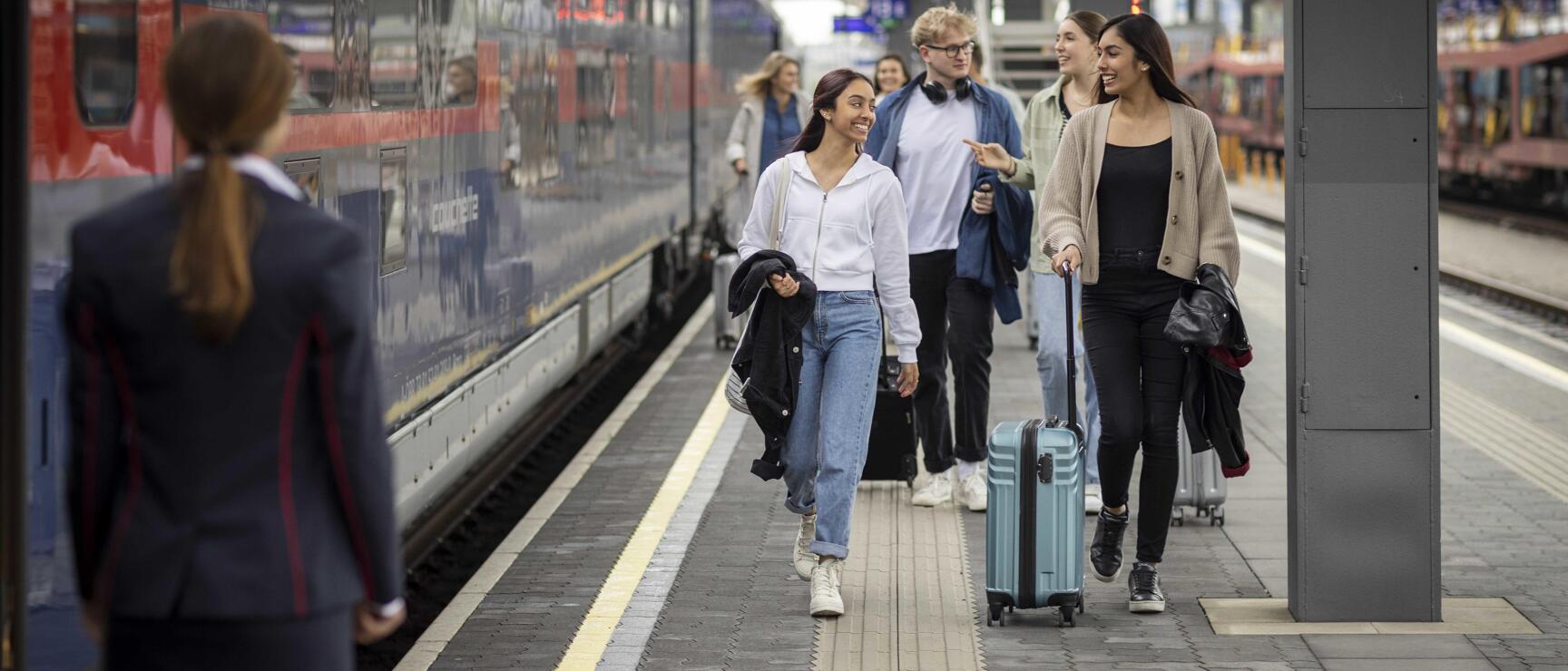 Young traveller with suitcase on platform beside blue night train, train attendant in foreground.