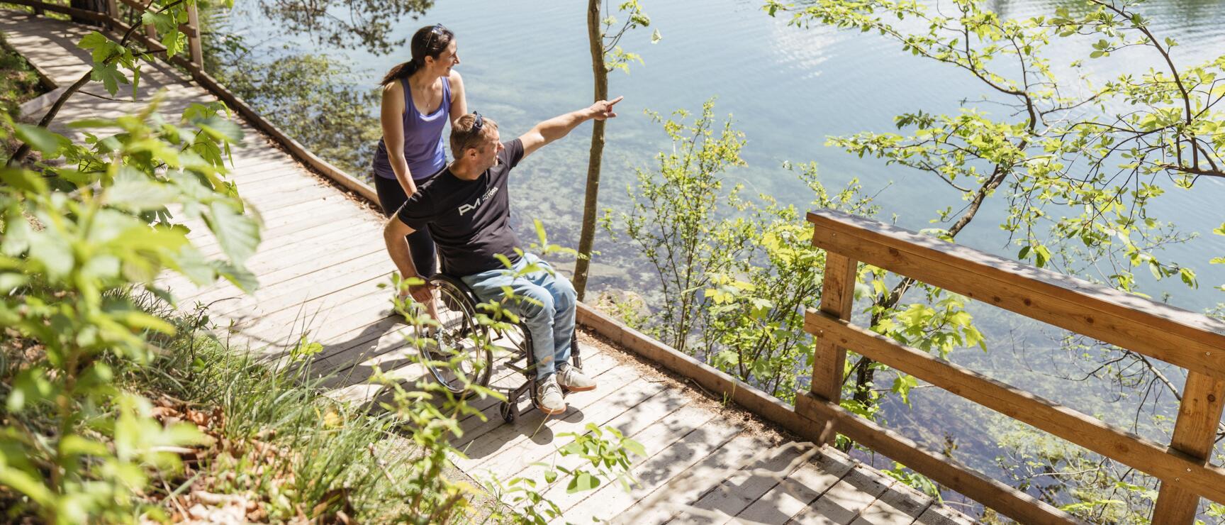 Wheelchair user pointing at a lake, woman standing behind, both on wooden dock, forest in background.