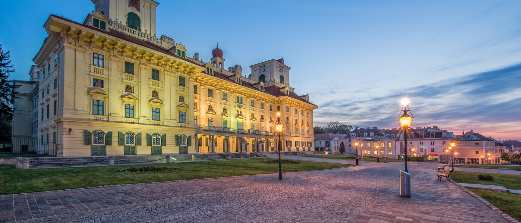 Esterházy Palace in Eisenstadt at dusk, illuminated, cobblestone square with street lamps in foreground.