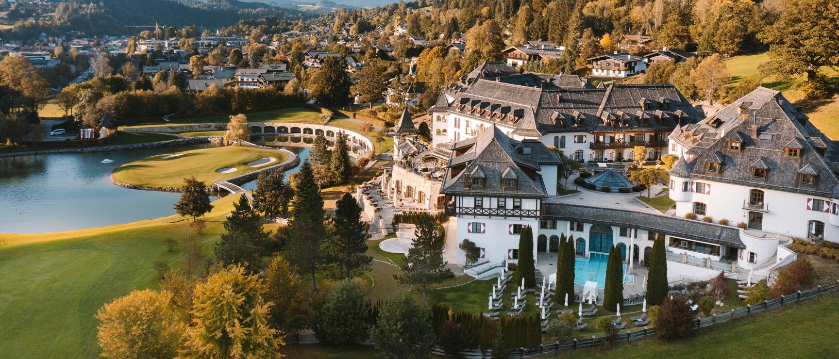 Aerial view of a white hotel complex with pool and golf course, lake and mountain panorama in autumn.