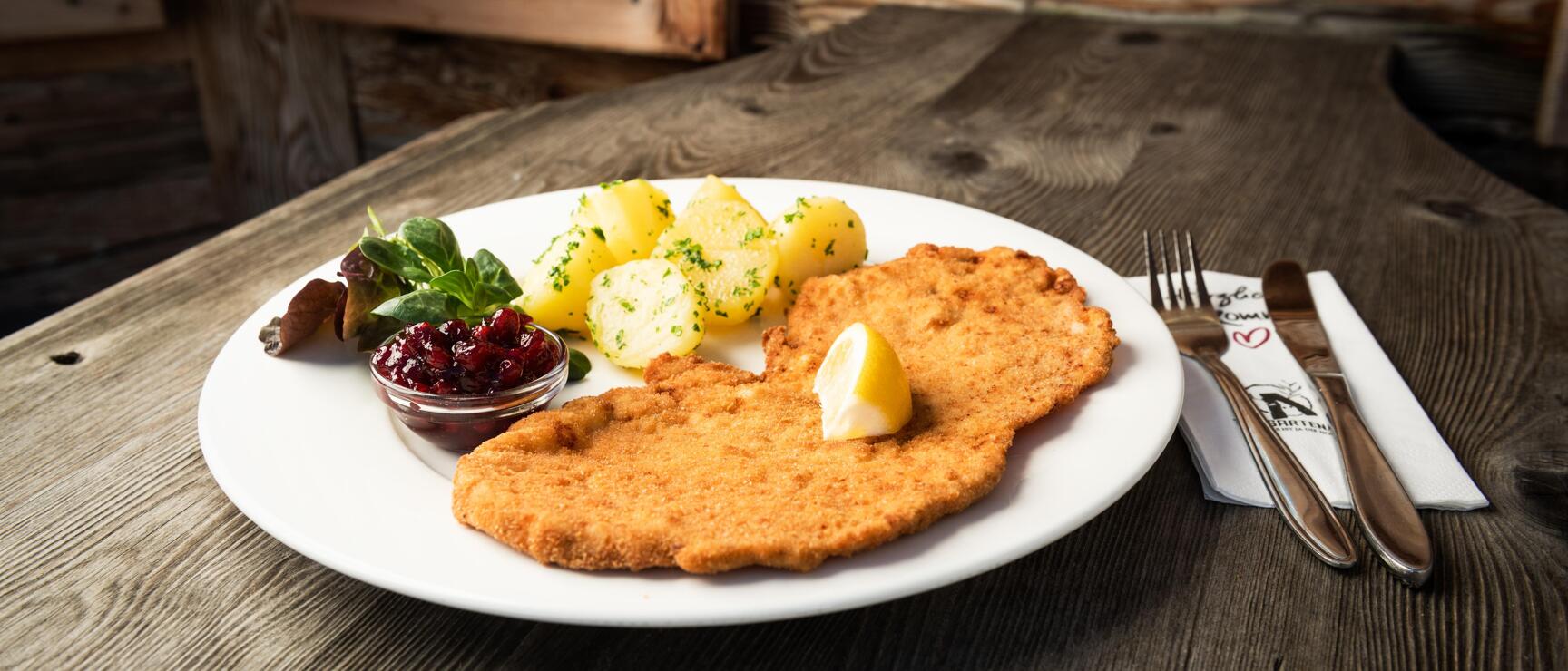 Wiener Schnitzel with lemon, parsley potatoes and lingonberries on white plate, wooden table.