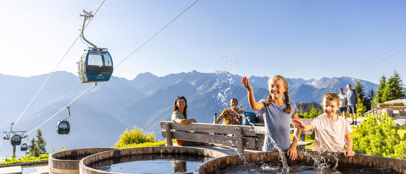 Two children splashing in wooden tubs at a mountain station, cable car and alpine panorama in the background.
