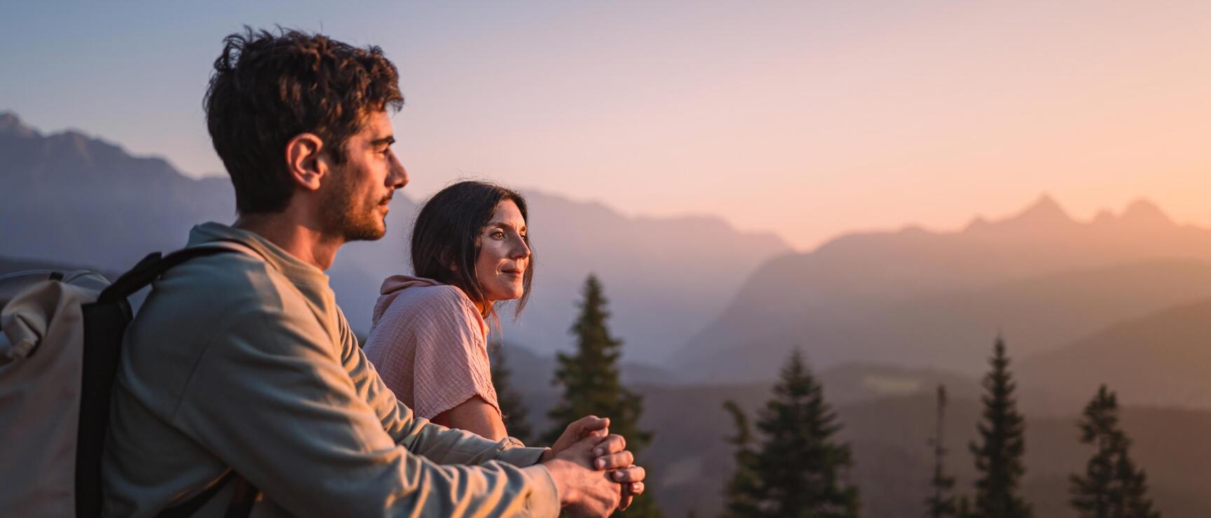 Two hikers with backpacks looking over mountain panorama with fir trees at sunset.