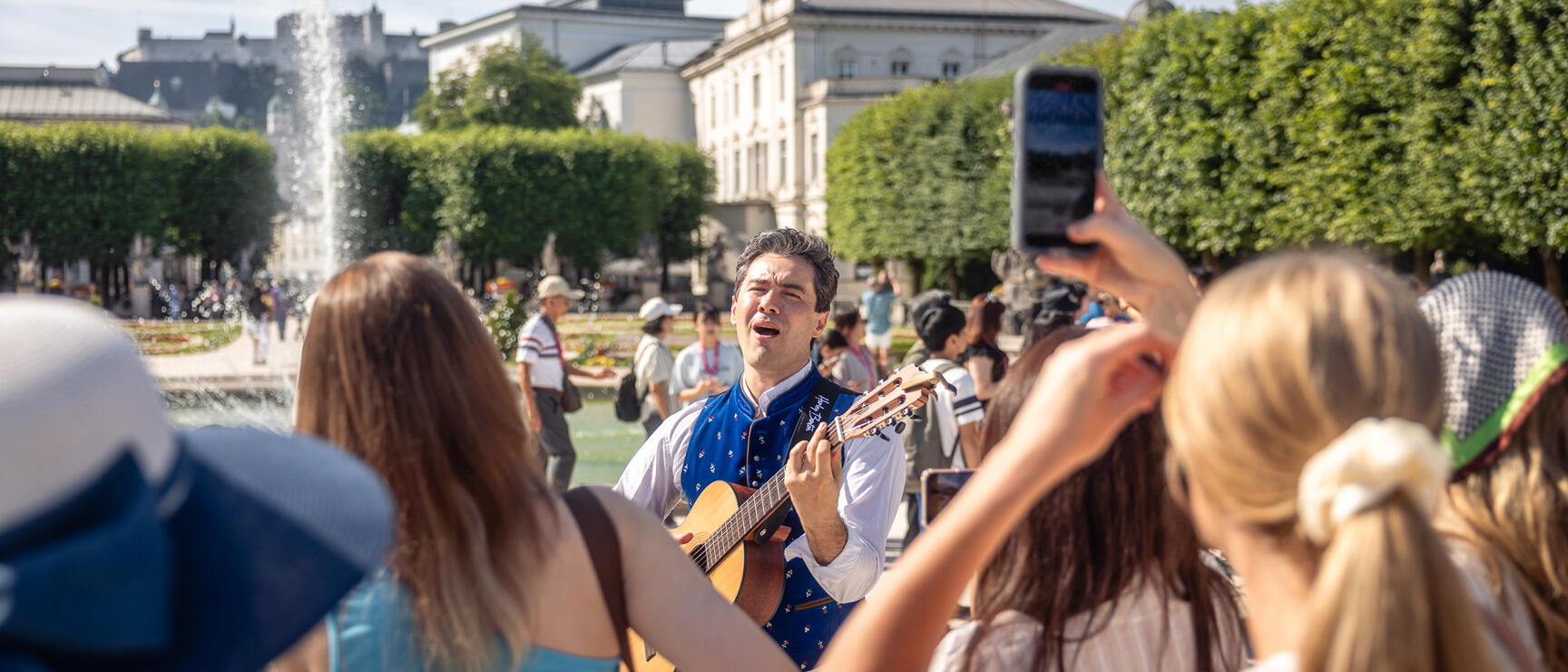 Guitarist in blue traditional costume sings in Mirabell Garden Salzburg, visitors filming him, fortress in the background.