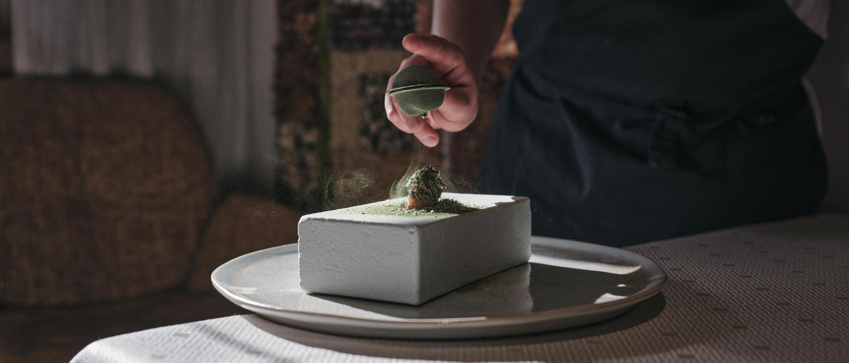 Chef in 'Das Karwendel' apron dusting green powder over angular dish on ceramic plate.