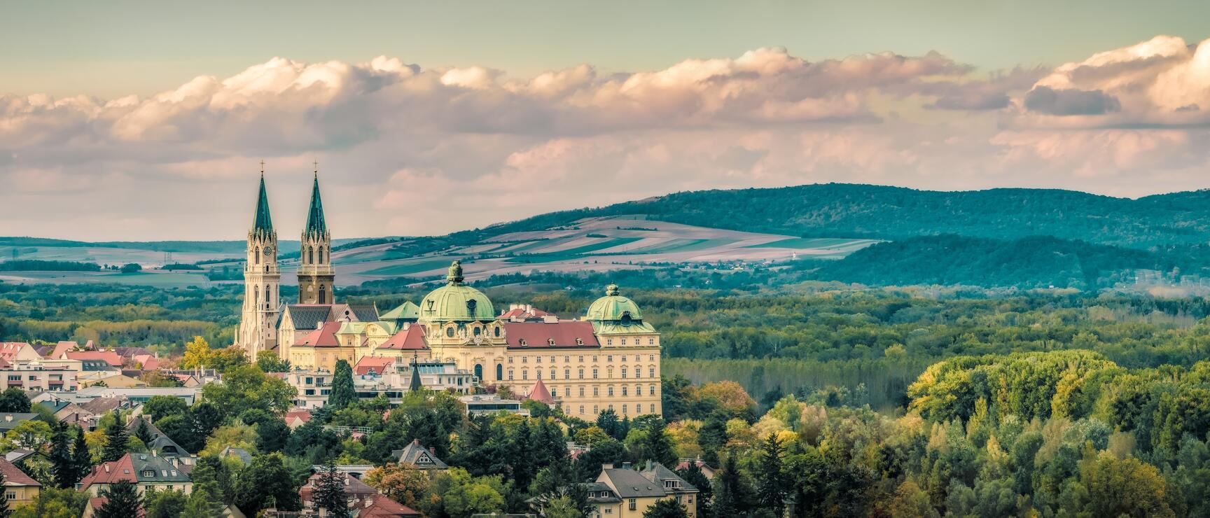 Panoramic view of Klosterneuburg with abbey, two church towers and green hilly landscape in background.