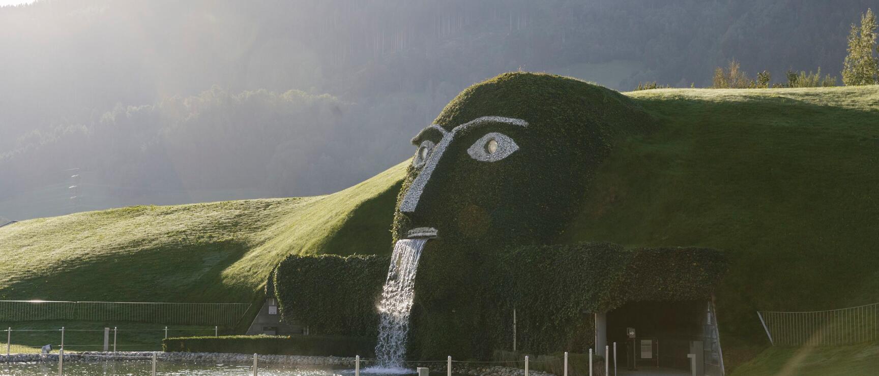 Giant head sculpture with waterfall at Swarovski Crystal Worlds in Wattens, pond and mountain backdrop.