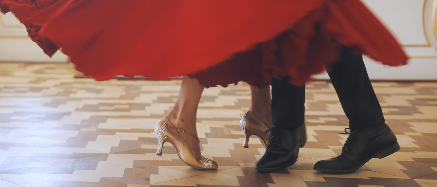 Dancing legs on parquet floor: red swirling dress, golden women's shoes, black men's shoes, hall in background.