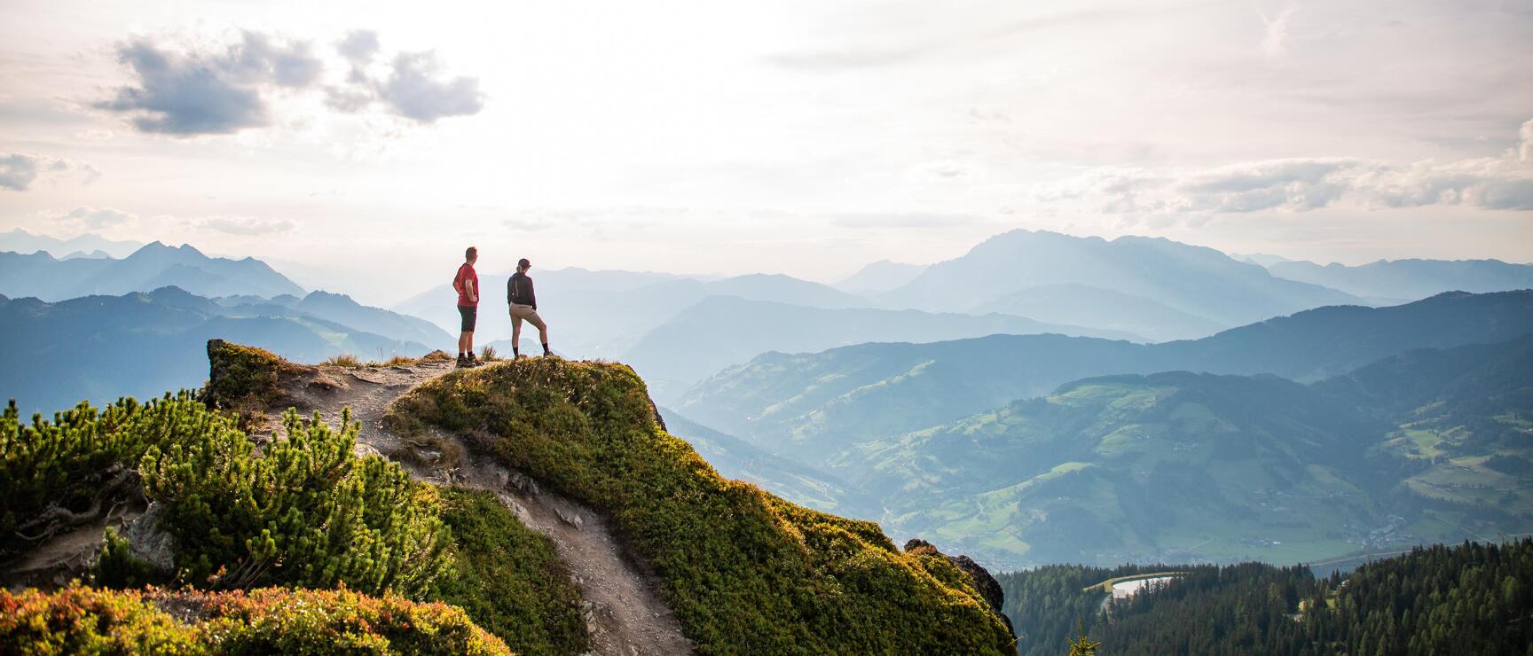 Two hikers on a rocky ridge with panoramic views of alpine valleys and mountain ranges.