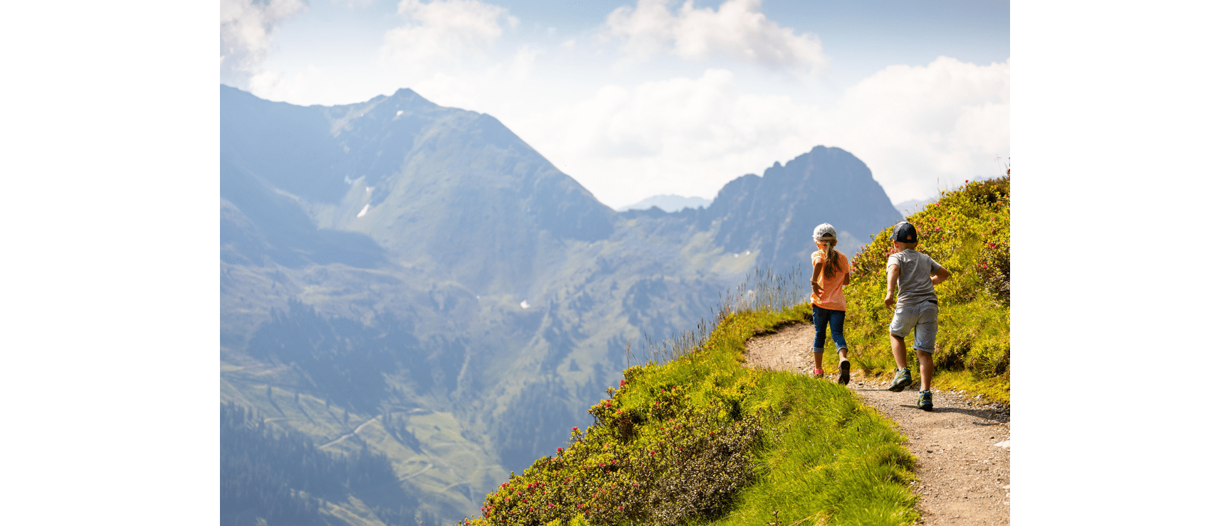 Two children running on an alpine path with alpine roses, wide valley and mountain peaks in the background.