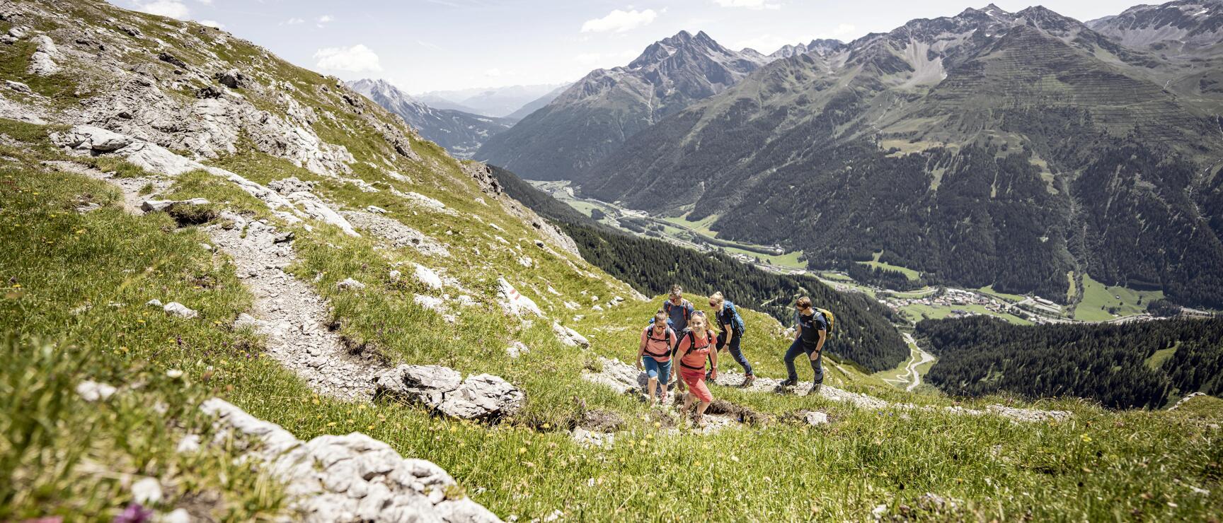 Five hikers on rocky mountain path, green valley with village and mountain range in background.