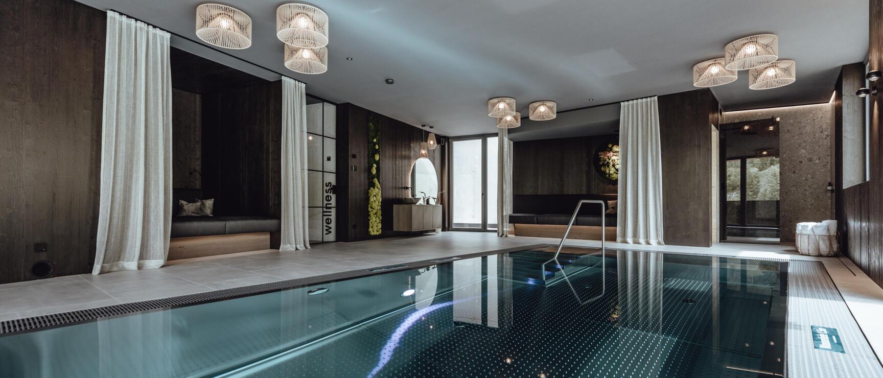 Indoor pool with dark mosaic tiles, white curtains and hanging lamps in a hotel wellness area.