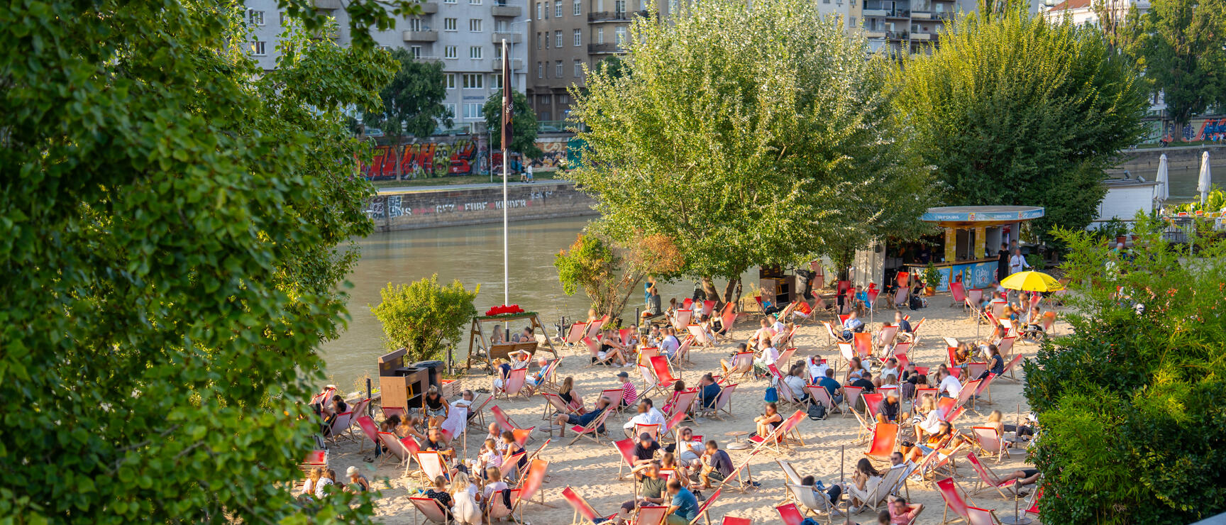 Many people in red deck chairs on a sandy beach at Danube Canal, Vienna, with trees and city buildings in background.