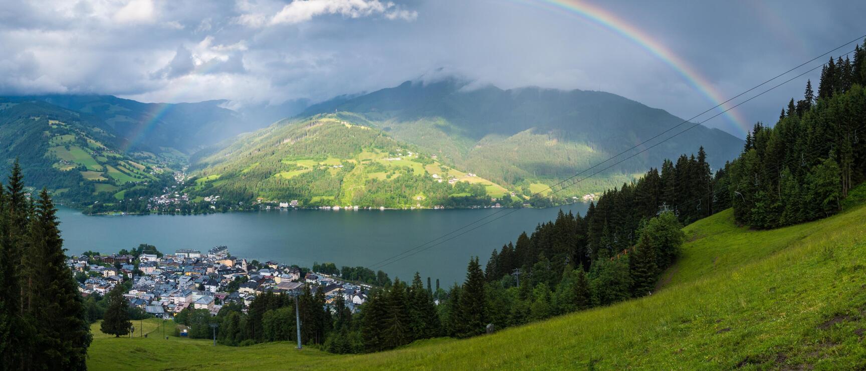 Panoramic view of Zell am See with Zeller See lake, green mountains and rainbow in cloudy sky.
