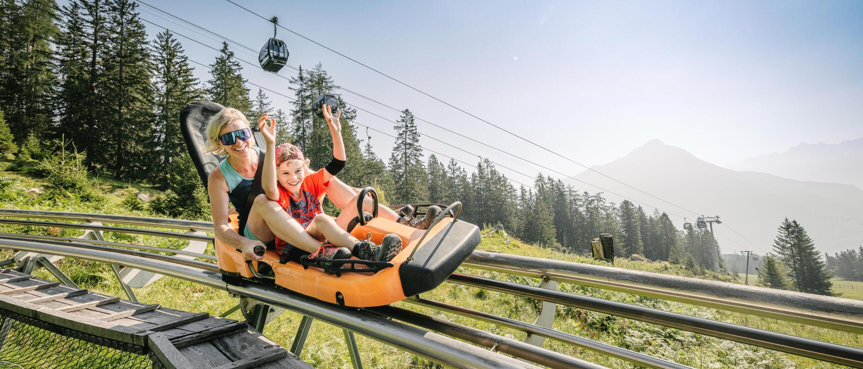 Frau und Kind in orangem Wagen auf einer Sommerrodelbahn, Gondelbahn und Berge im Hintergrund.